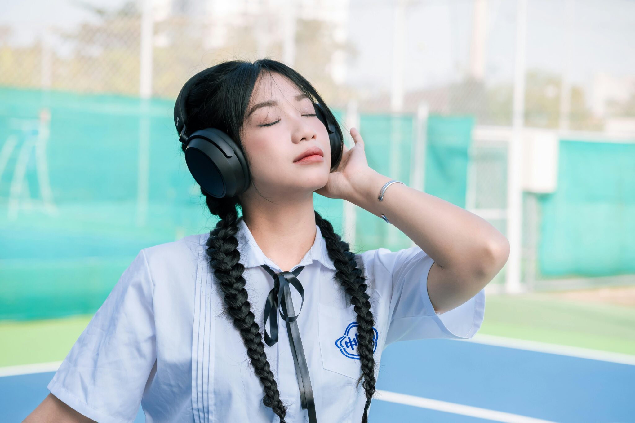 Asian girl in school uniform with braided hair listens to music outdoors.